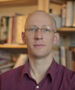 Portrait of a white male guy, with shaved head, blue eyes, with glasses, in front of a library